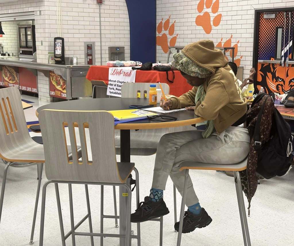 A student is sitting at a table in a school cafeteria writing on a notebook.