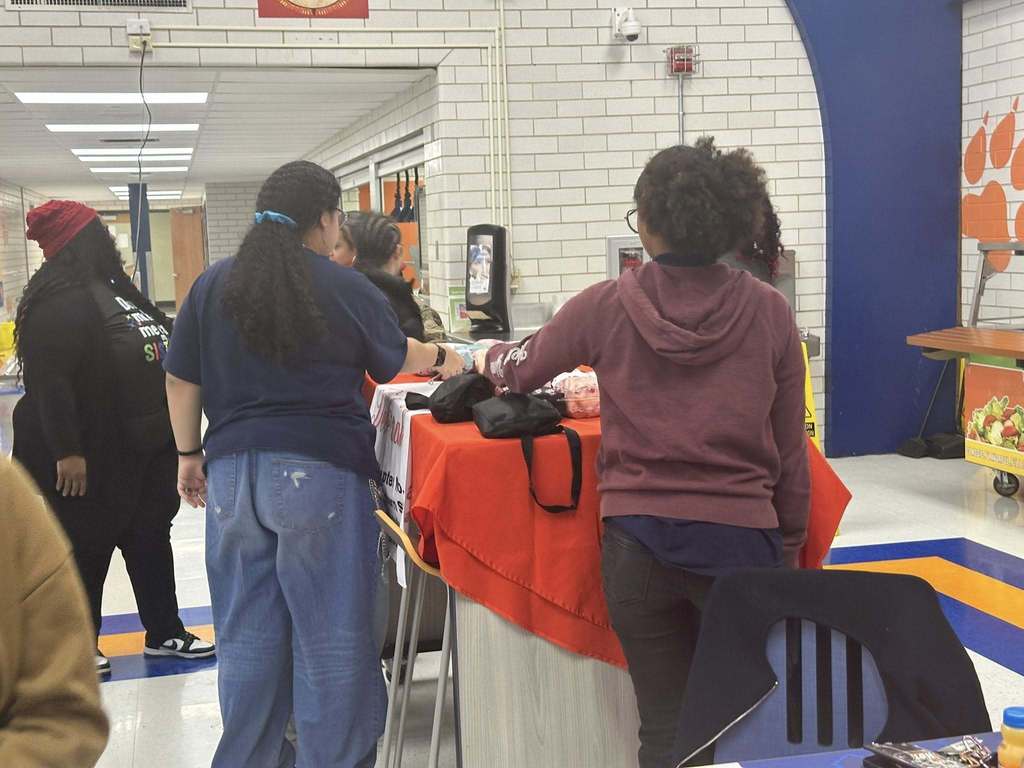 Two students are reaching for pouches that are on a table in a school cafeteria. Other people can be seen in the background.