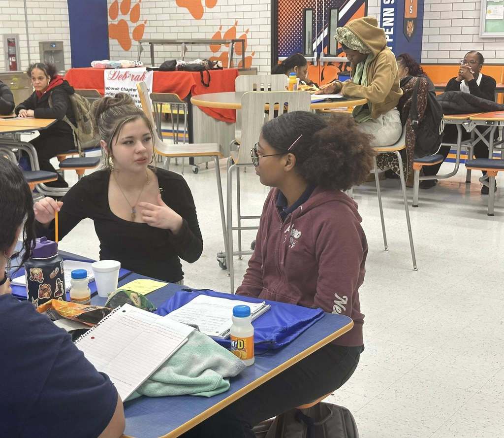Two students are sitting at a table in a school cafeteria, engaging in a conversation. Other people can be seen nearby.