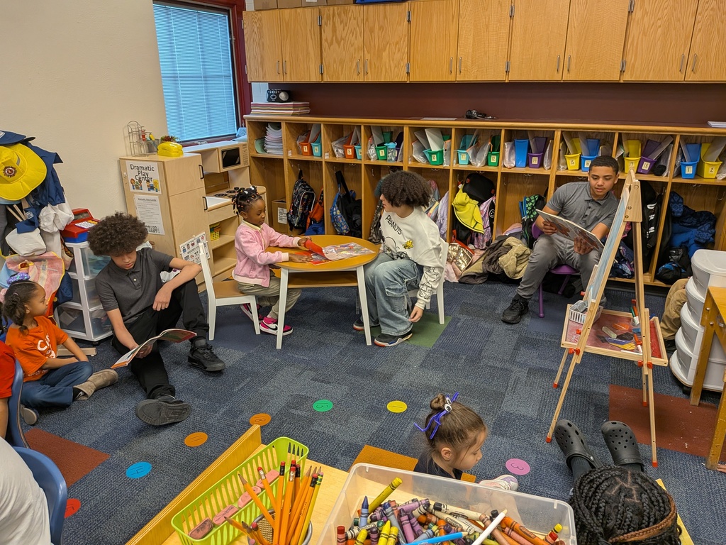 A diverse group of students are seated around a table and on the floor in a school classroom, engaging in reading colorful picture books.