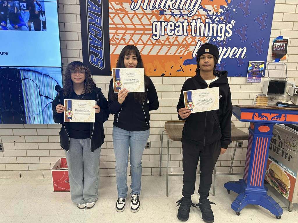 Three students are holding certificates while standing in front of a school-themed mural with positive messages in a school cafeteria.