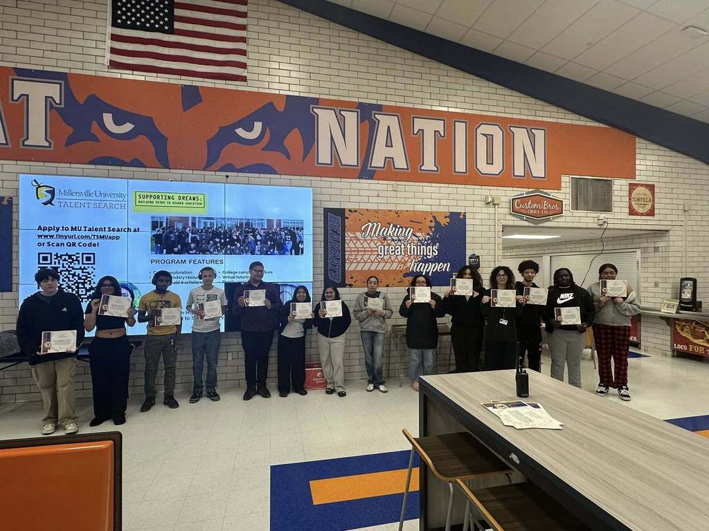 A group of students are holding certificates in a school cafeteria, while standing in front of a screen that has information about a university talent search program.