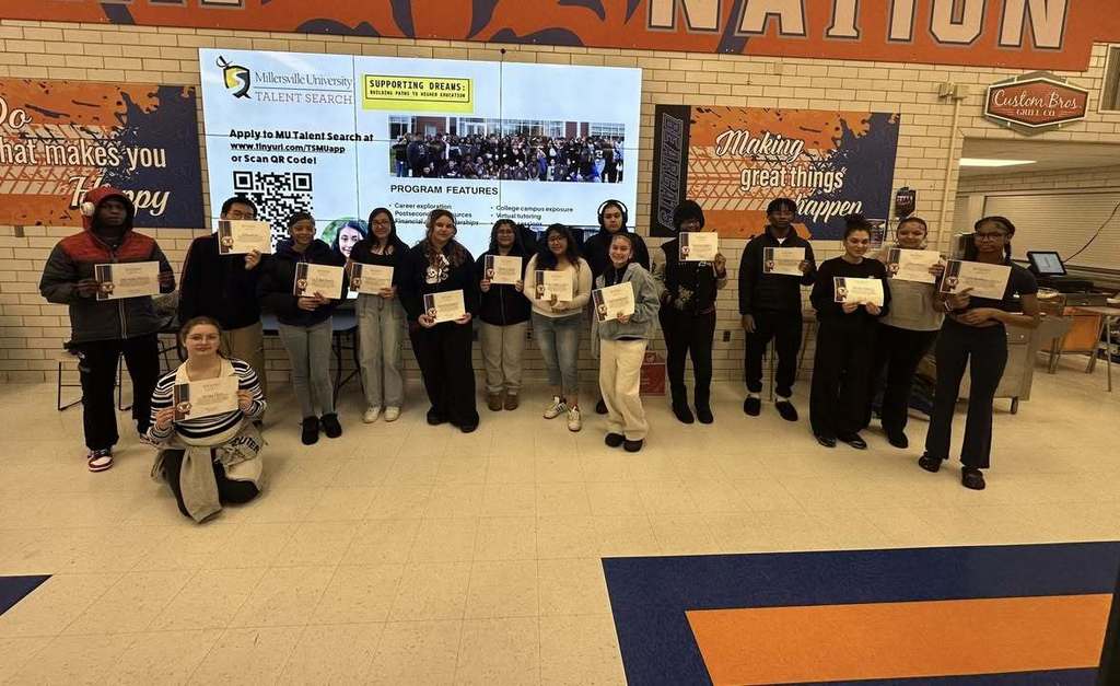 A group of students are holding certificates in a school cafeteria, while standing in front of a screen that has information about a university talent search program. One of the students is kneeling down on the floor.