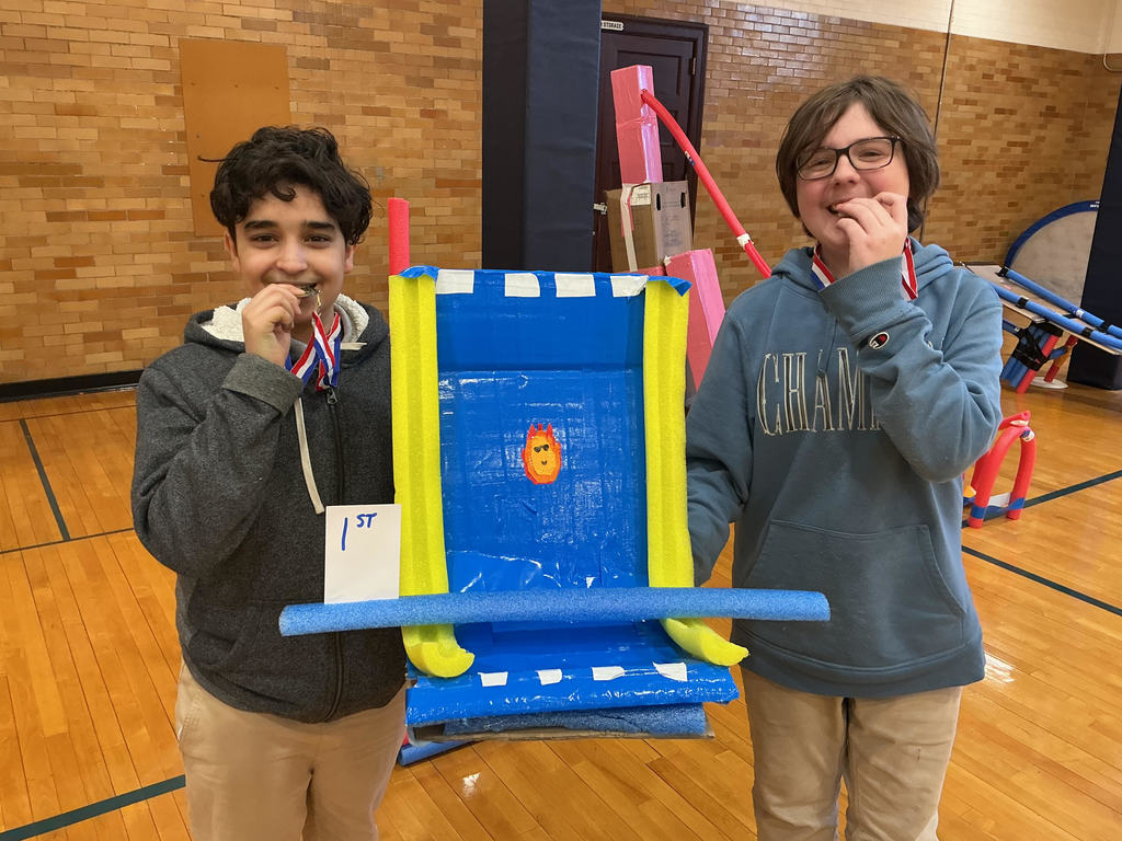 Two students are standing in a school gym wearing medals and holding a handmade boat made of blue and yellow materials.  