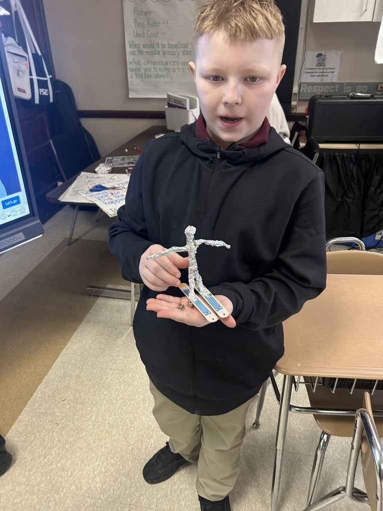 A student is standing near a desk in a school classroom, holding a small sculpture made of foil and popsicle sticks.