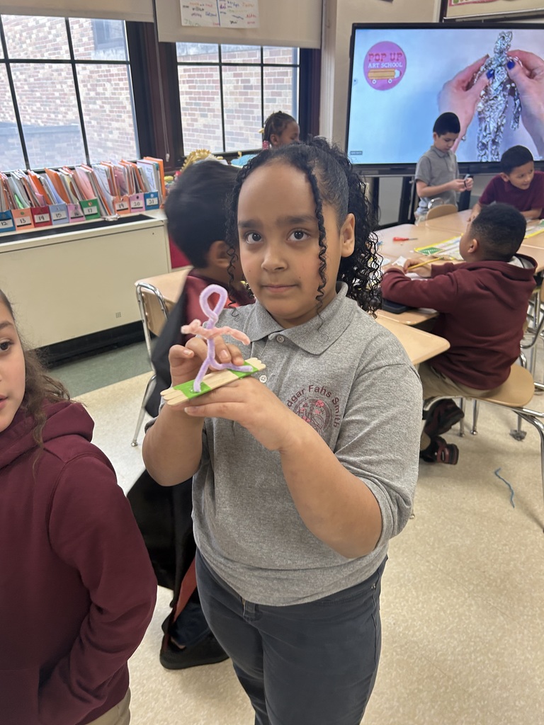 A student is standing in a school classroom, holding a small art project made from pipe cleaners and paper. Other students can be seen nearby in the background.