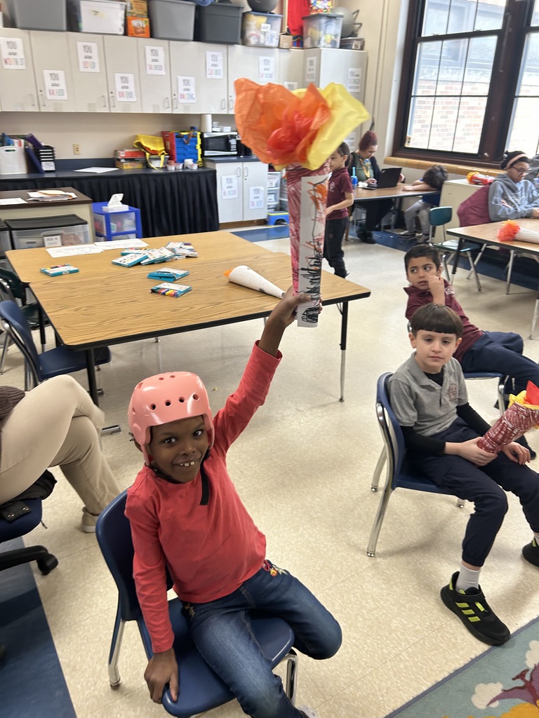 A student wearing a pink helmet can be seen siting on a chair, raising a paper torch with red and yellow tissue flames in the air in a school classroom. Other students can be seen sitting in chairs nearby.