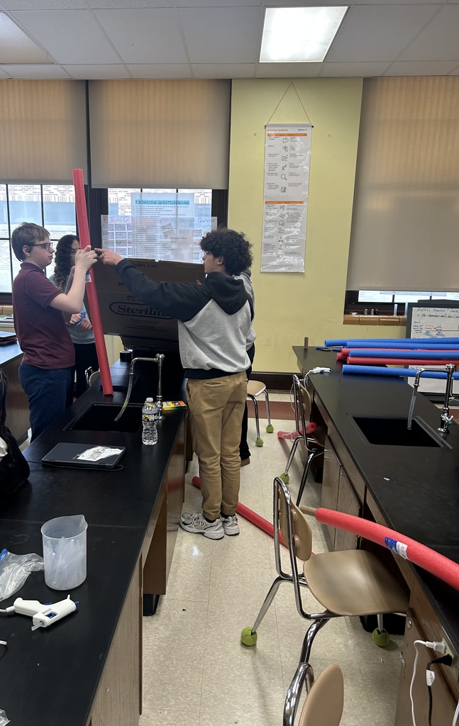 A group of students are collaborating on a project in a school classroom. Red and blue pool noodles can be seen nearby.