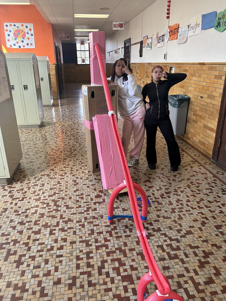Two students are standing in a school hallway, surrounded by lockers and art-covered walls. A tall cardboard and pool noodle structure is being displayed.