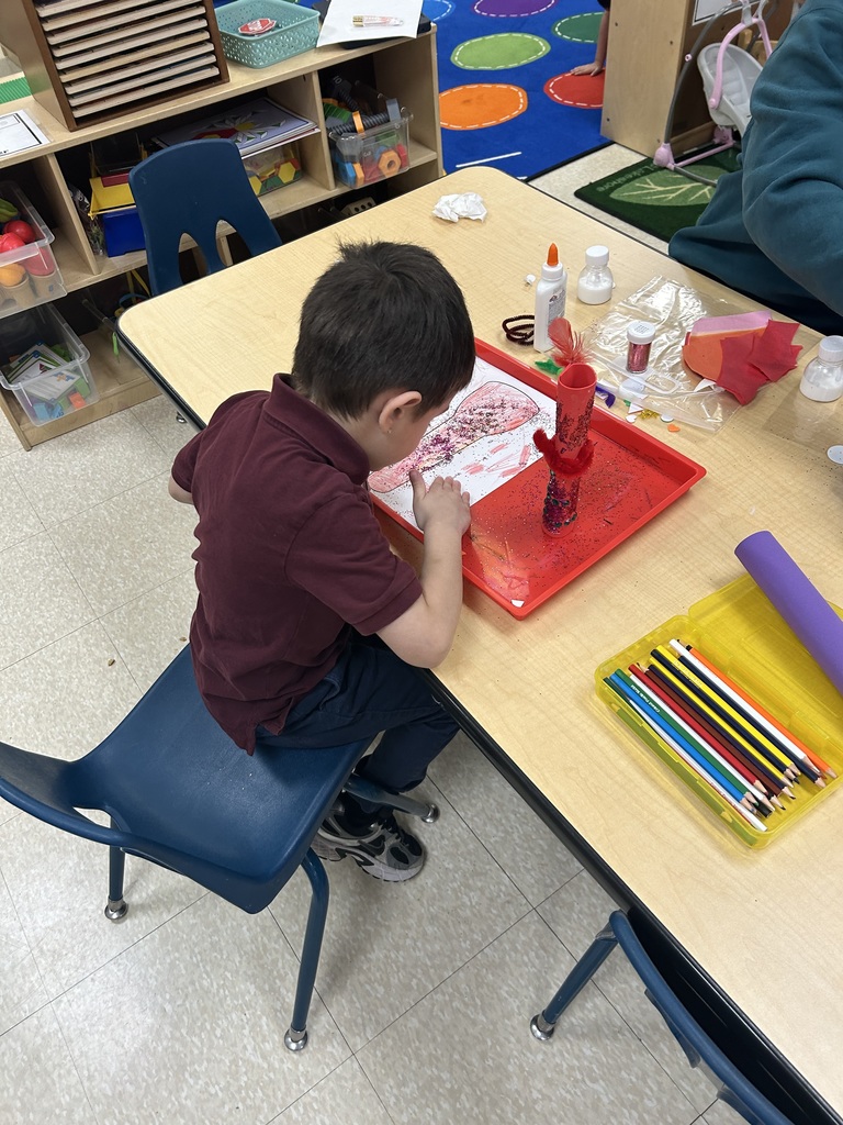 A student is sitting at a table in a school classroom, engaging in an art activity using red and black glitter. Other craft supplies can be seen on the table.