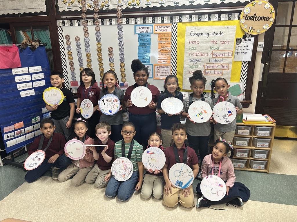 A group of students are sitting and standing in front of a wall in a school classroom, holding decorated paper plates. Educational posters can be seen on the wall.