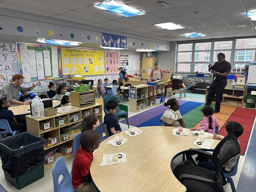 A group of students are sitting at tables in a school classroom, listening to a district staff member who is standing at the front of the classroom. Other district staff members can be seen supervising nearby.