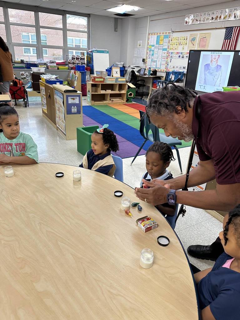 A district staff member is helping a student who is sitting at a table in a school classroom. Other students can be seen observing at the table. The students are engaging in an activity using small jars. Educational materials can be seen in the background.