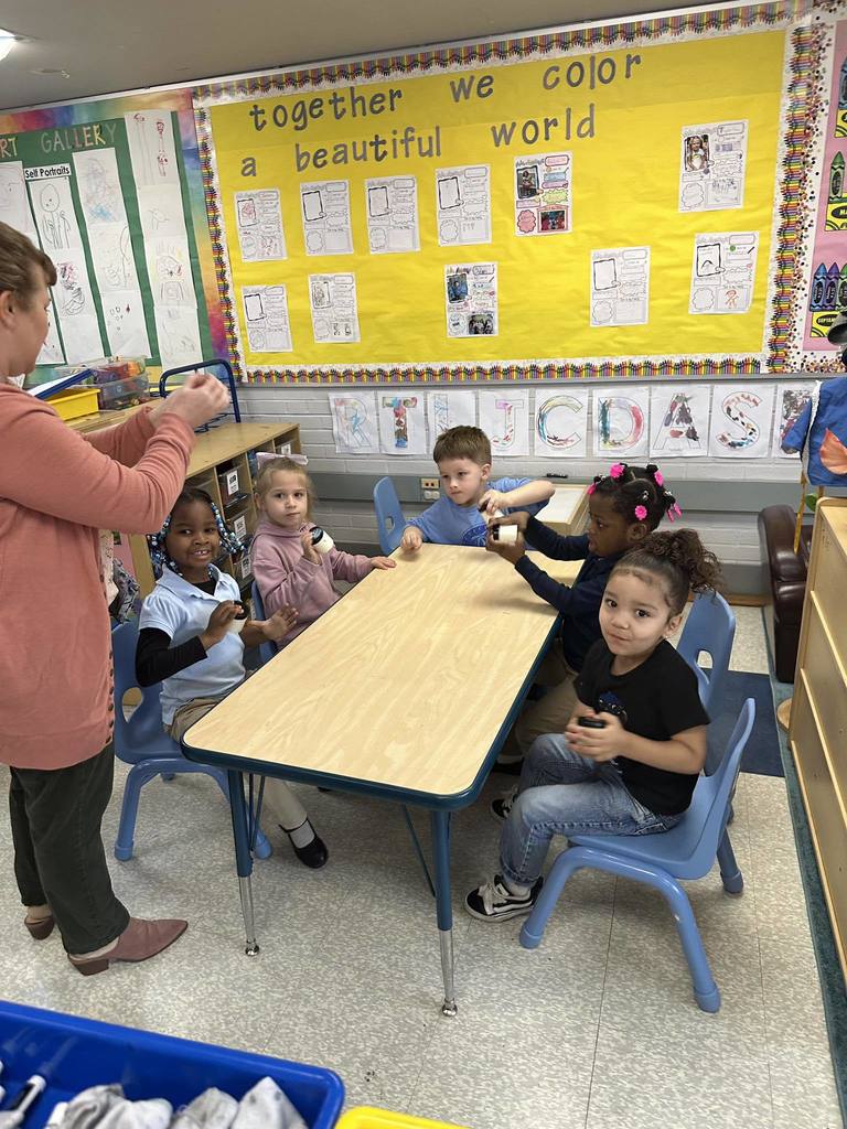 A district staff member is standing near a group of students who are sitting at a table in a school classroom. The students are holding and shaking jars in their hands. Educational materials can be seen in the background.