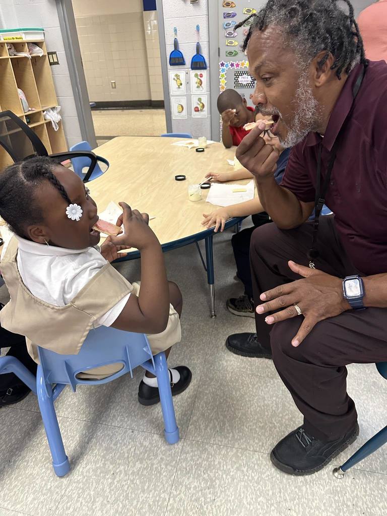 A district staff member and a student are mimicking eating gestures in a school classroom. Other students can be seen nearby in the background.