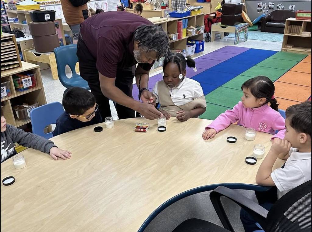 A district staff member is helping a student who is sitting at a table in a school classroom. Other students can be seen observing at the table. The students are engaging in an activity using small jars. Educational materials can be seen in the background.