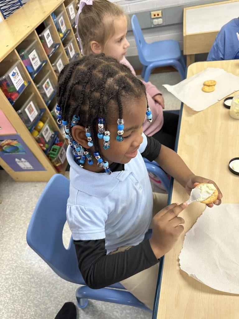 Two students are sitting at a table in a school classroom. One of the students is spreading cream on a cracker. 