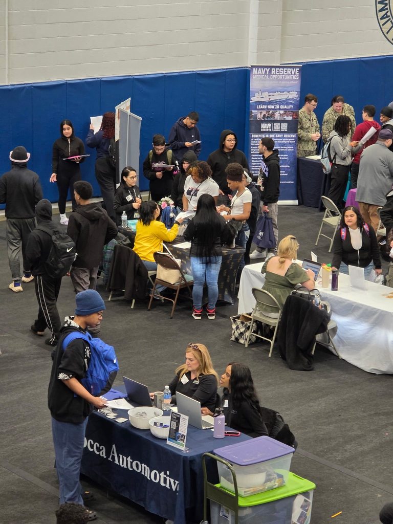A bustling college fair inside a school gymnasium filled with students and representatives at booths with colorful tablecloths, engaging in conversations.