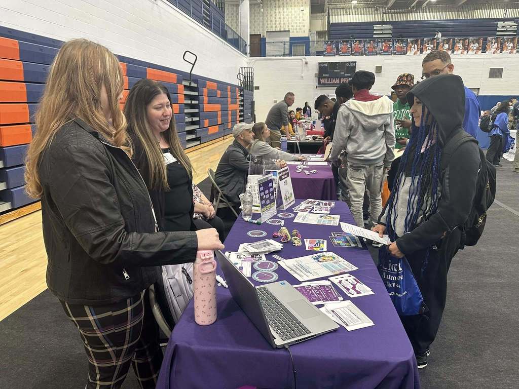 A bustling college fair inside a school gymnasium filled with students and representatives at booths with colorful tablecloths, engaging in conversations.