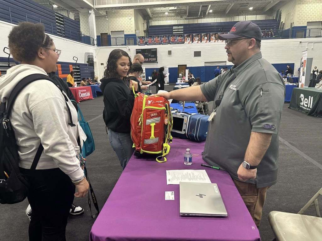 A bustling college fair inside a school gymnasium filled with students and representatives at booths with colorful tablecloths, engaging in conversations.