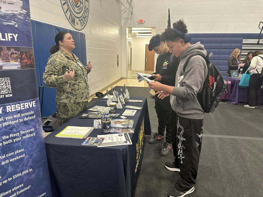 A bustling college fair inside a school gymnasium filled with students and representatives at booths with colorful tablecloths, engaging in conversations.