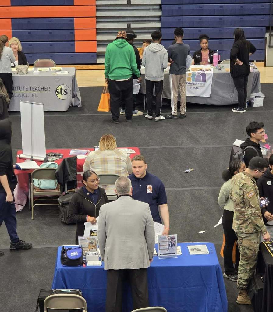 A bustling college fair inside a school gymnasium filled with students and representatives at booths with colorful tablecloths, engaging in conversations.