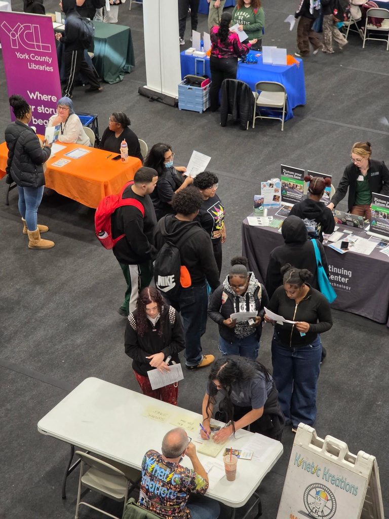 A bustling college fair inside a school gymnasium filled with students and representatives at booths with colorful tablecloths, engaging in conversations.