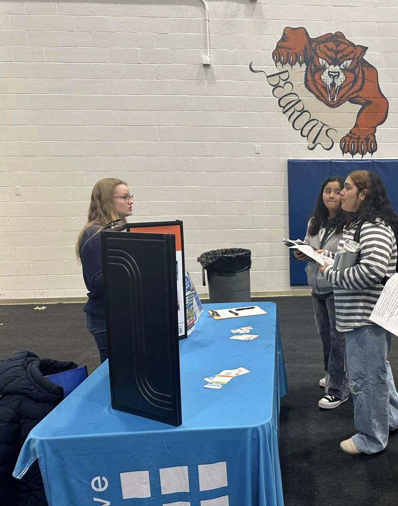 A bustling college fair inside a school gymnasium filled with students and representatives at booths with colorful tablecloths, engaging in conversations.