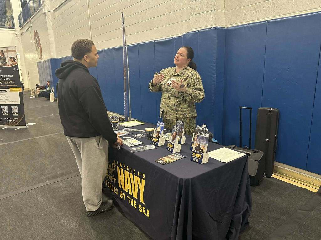 A bustling college fair inside a school gymnasium filled with students and representatives at booths with colorful tablecloths, engaging in conversations.