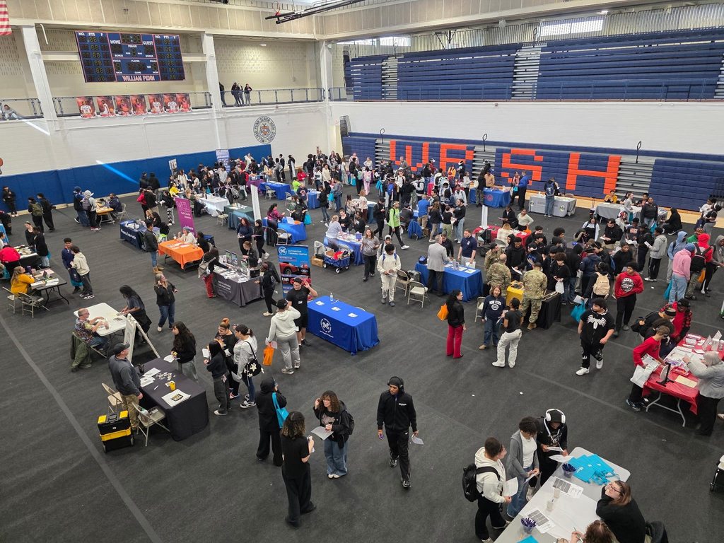 A bustling college fair inside a school gymnasium filled with students and representatives at booths with colorful tablecloths, engaging in conversations.