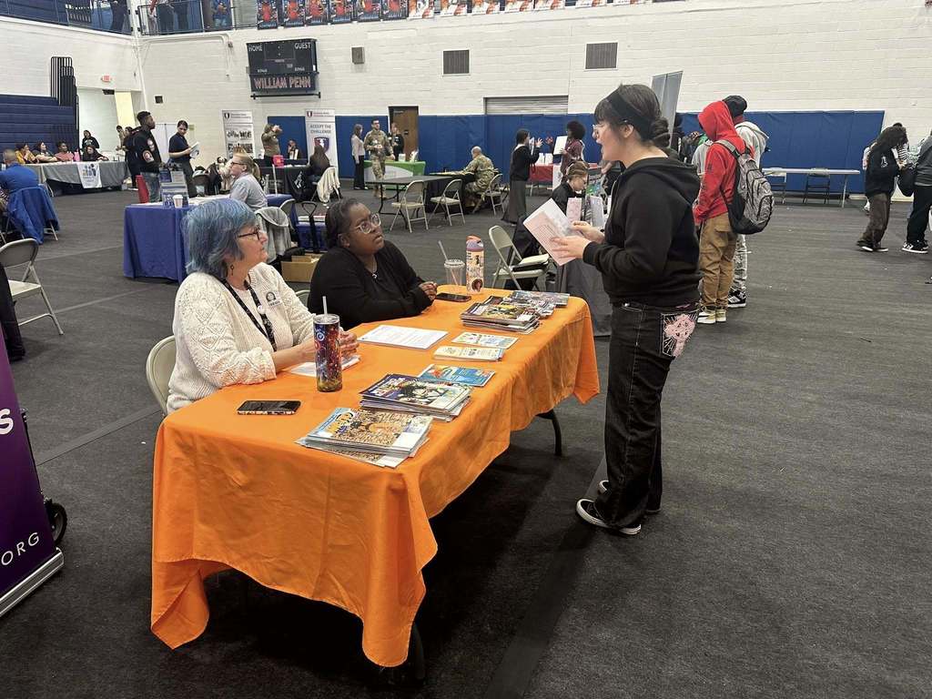 A bustling college fair inside a school gymnasium filled with students and representatives at booths with colorful tablecloths, engaging in conversations.