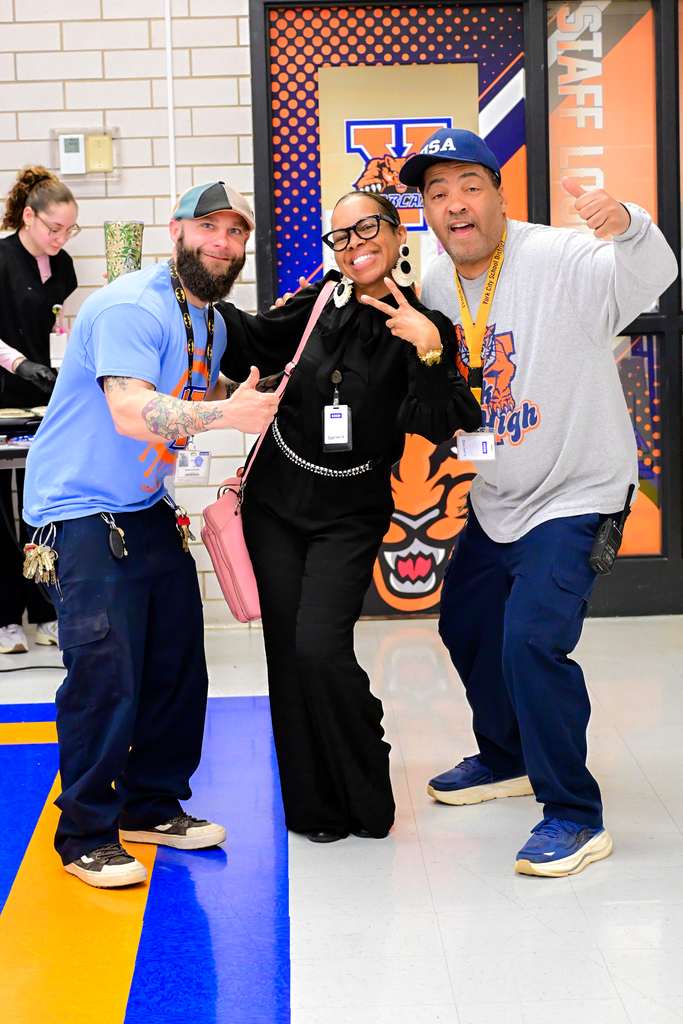 Three district staff members are posing together in a school cafeteria. Another district staff member can be seen nearby in the background.