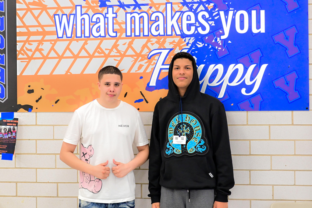 Two students are standing together in front of a wall in a school cafeteria.