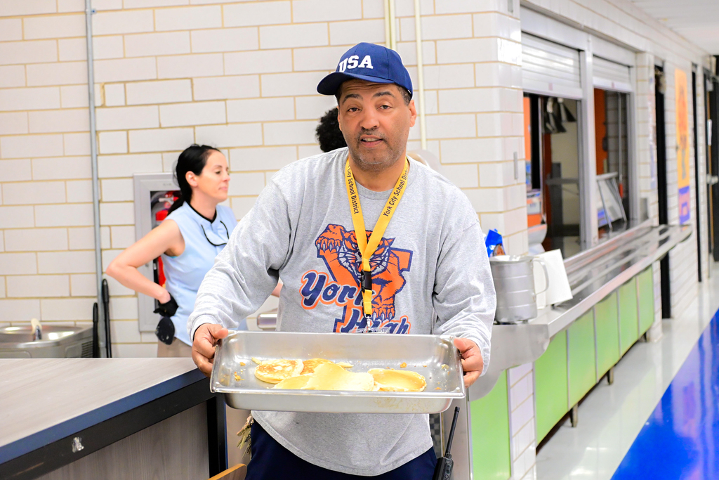 A district staff member is holding a tray of pancakes while standing in a school cafeteria. Another district staff member can be seen nearby in the background.