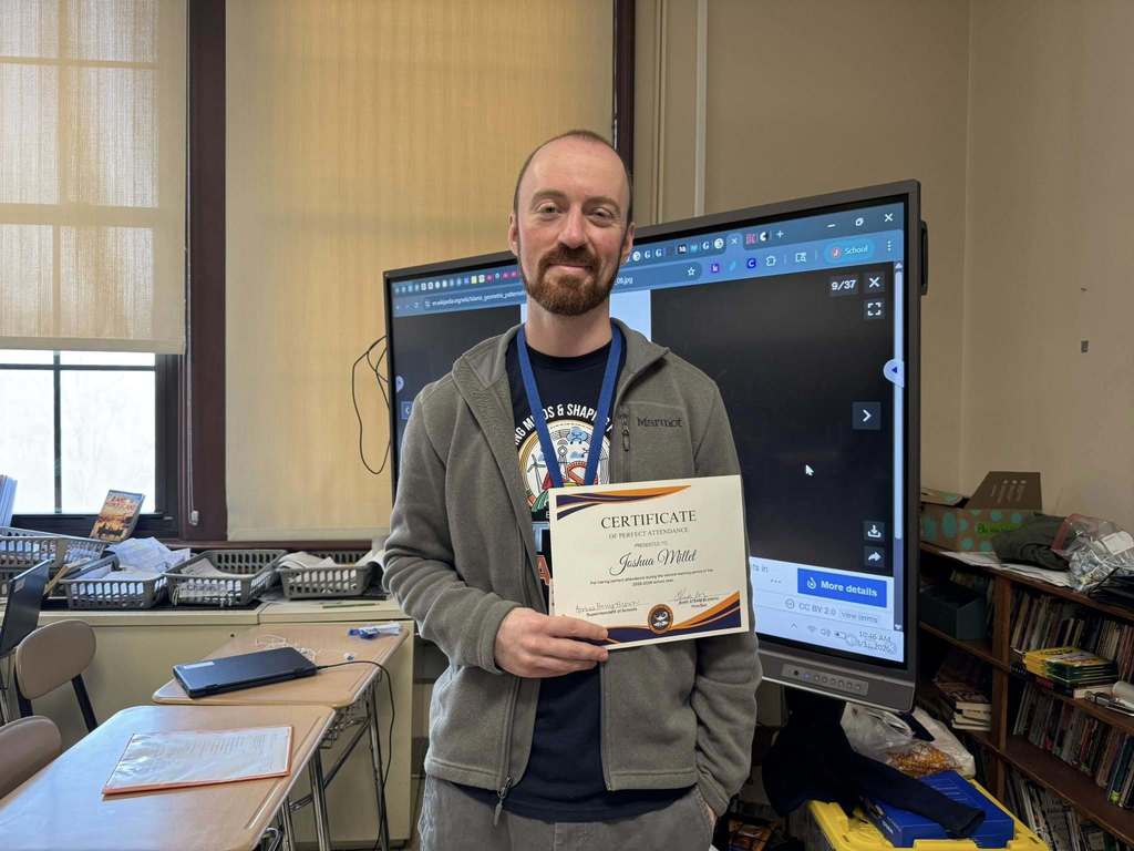 A district staff member is standing near a large screen, holding a Perfect Attendance certificate for the second marking period.