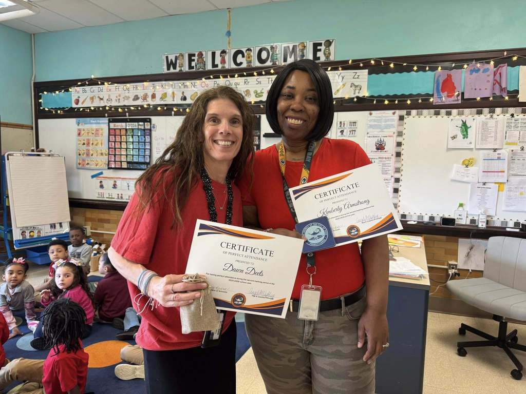 Two district staff members are standing in a school classroom, both of them holding Perfect Attendance certificates for the second marking period.