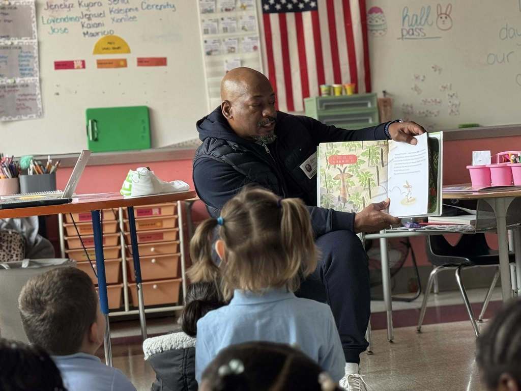 A local male leader is reading a book to a group of young students who are sitting down in a school classroom. 
