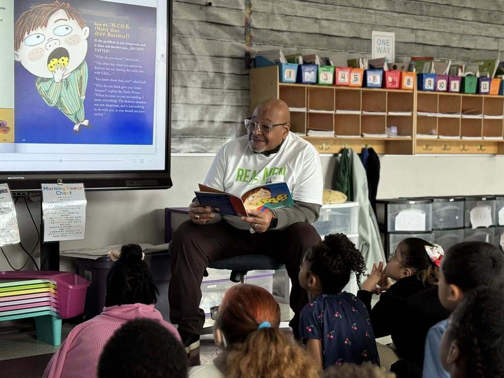 A local male leader is reading a book to a group of young students who are sitting down in a school classroom. 