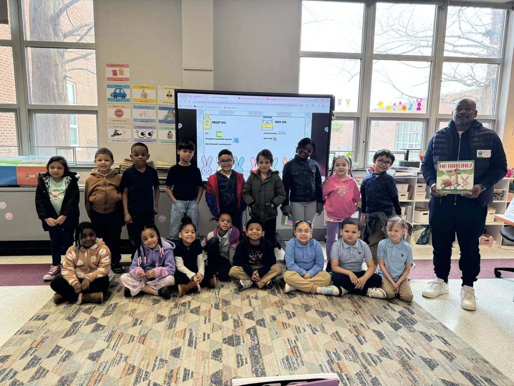 A local male leader is standing next to a group of young students in a school classroom. 