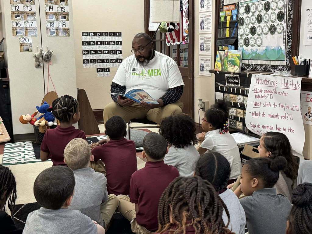 A local male leader is reading a book to a group of young students who are sitting down in a school classroom. 