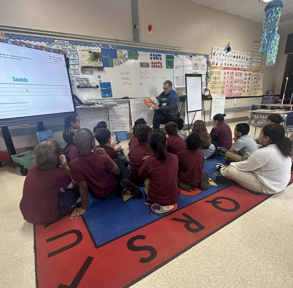 A local male leader is reading a book to a group of young students who are sitting on a colorful carpet in a school classroom. 