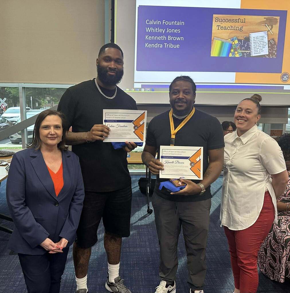 District administrators and staff members are posing inside of a conference room. Both of the staff members are holding certificates for Awards of Appreciation.