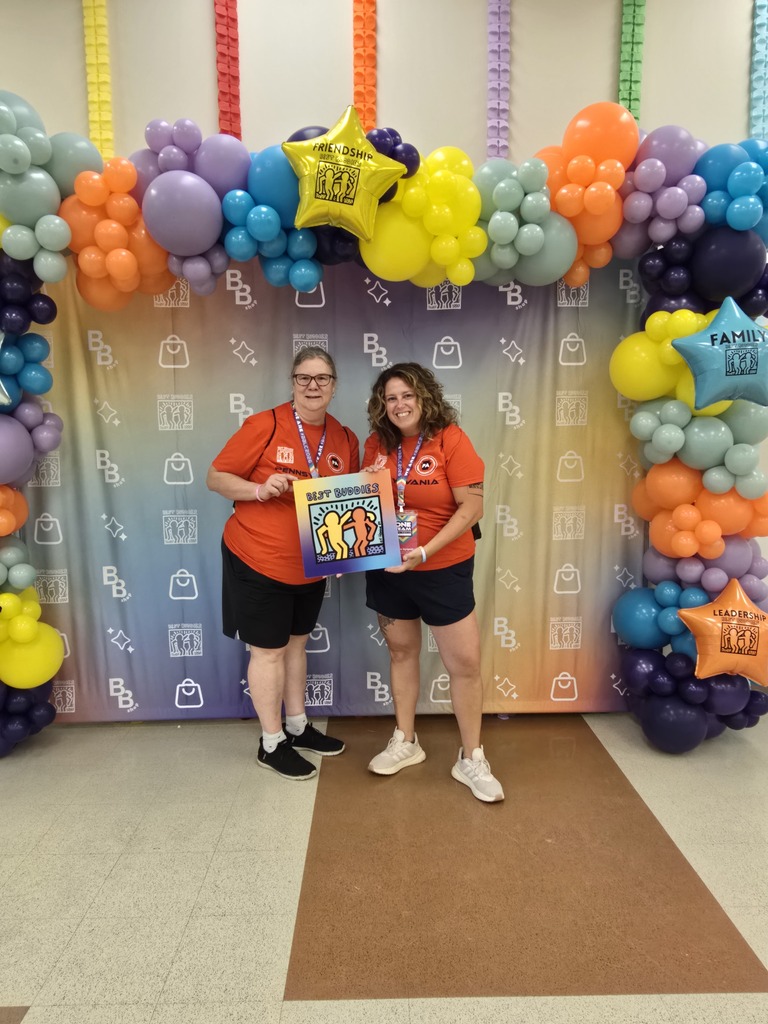 Two district staff members are posing together with a Best Buddies sign beneath a colorful balloon arch at a Best Buddies Leadership Conference.