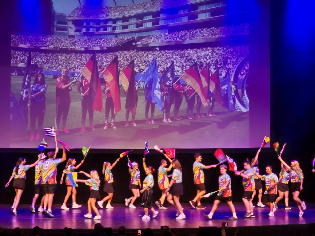 Best Buddies participants are waving international flags during a colorful opening ceremony performance on stage.