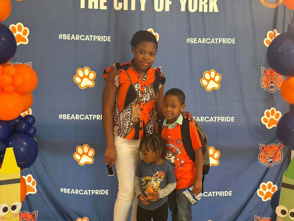 A district family consisting of a woman and two young children, are posing in front of a blue Bearcat Pride school backdrop decorated with orange and navy balloons and paw print logos.