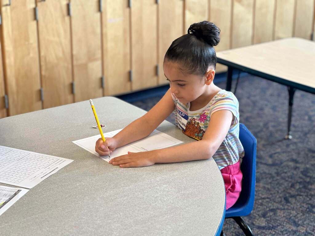 A young student is sitting at a classroom table, focused on writing with a pencil on a worksheet.