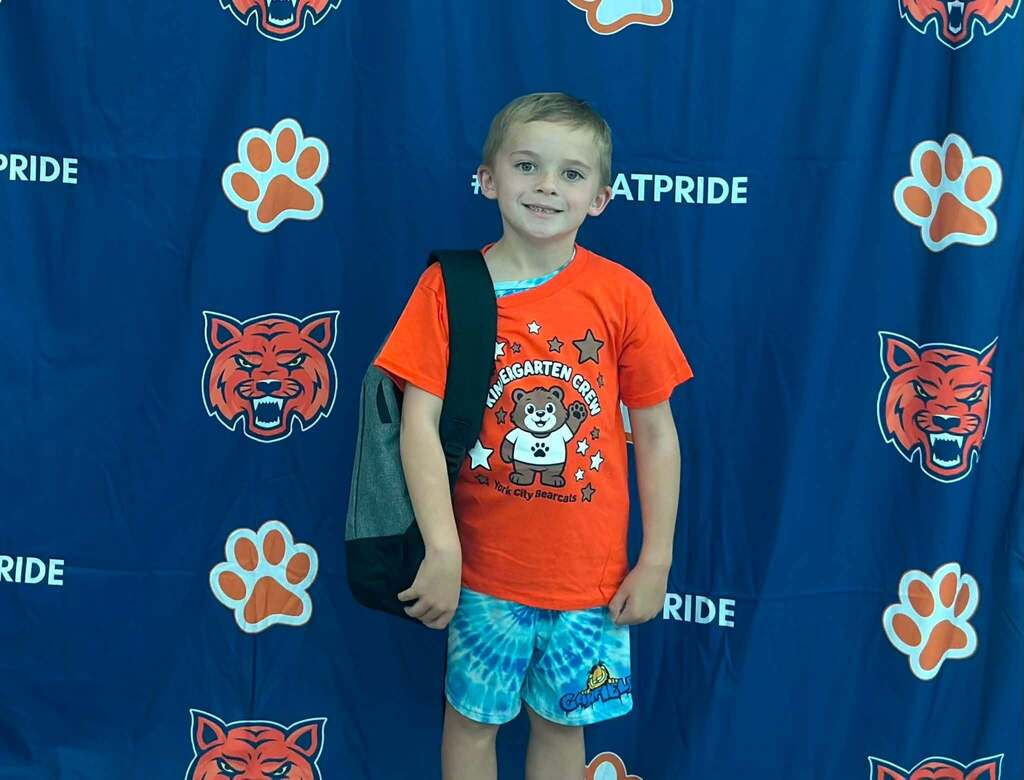 A young student wearing an orange Kindergarten Crew shirt and backpack is posing in front of a Bearcat Pride backdrop during kindergarten orientation.