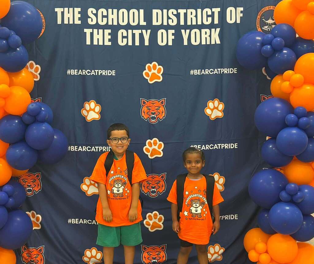 Two young students wearing bright orange Kindergarten Crew shirts and backpacks are standing in front of a School District of the City of York Bearcat Pride backdrop decorated with blue and orange balloon arches.