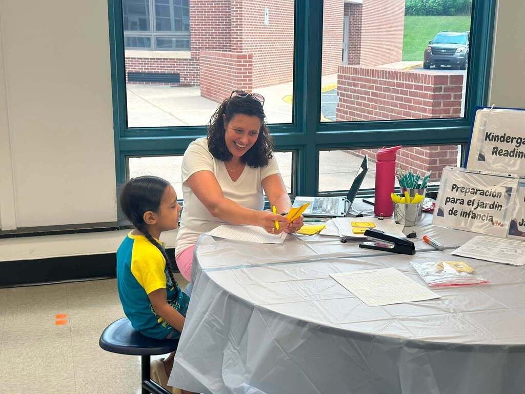 A district staff member is smiling while helping a young student at a kindergarten readiness registration table with school materials and bilingual information displayed.