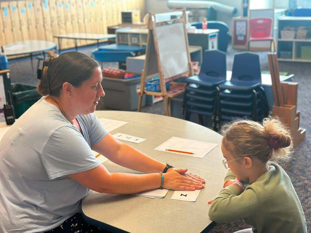 A district staff member is working one-on-one with a young student on a letter recognition activity at a classroom table.